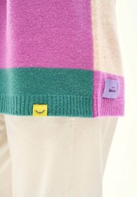 Dolná časť farebného bloku svetra v ružovej, zelenej a béžovej farbe s žltou nášivkou zobrazujúcou dizajn zatvoreného oka, nosená cez krémové nohavice.
