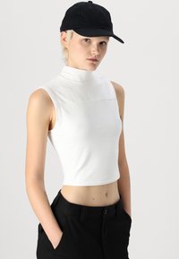 Bílý bezrukávový crop top s vysokým límcem a hladkým povrchem, v kombinaci s černými kalhotami, nošený s černou čepicí. Jednoduché pozadí.