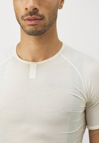 T-shirt de sport ajusté beige clair, à manches courtes, col rond et avec des lignes horizontales subtiles. Présente une petite étiquette blanche au col.