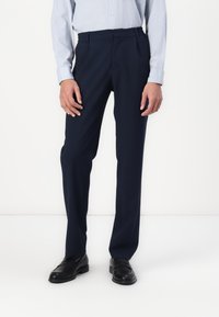 Marineblaue Hose aus glattem Stoff, mit tailliertem Schnitt, flachem Vorderteil und einer sauberen Bügelfalte entlang des Beins. Kombiniert mit schwarzen Slippern.