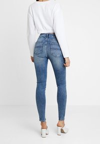 Jeans skinny de mezclilla azul con cintura de altura media, que cuentan con bolsillos traseros y un diseño ajustado. Combinados con una blusa blanca de manga larga corta.