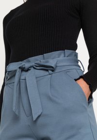 Blå högmidjade byxor med en bältesknut, tillverkade av ett slätt material. Har sidofickor och en diskret veckad design. Svart ribbad topp synlig.