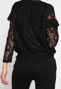 Blouse en dentelle noire avec des manches longues et transparentes ornées de motifs floraux, taille froncée et col rond. Design texturé et légèrement drapé.