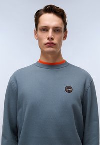Grå hettegenser med en rund logo, med oransje indre krage, rund hals og lange ermer. Glatt tekstur, uformelt design.