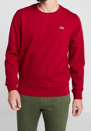 Mand iført en ensfarvet rød langærmet sweatshirt med en lille grøn krokodillelogo på brystet og grønne bukser.