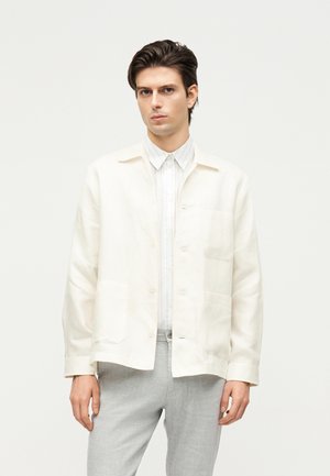 Fiatal férfi, aki törtfehér gombos ingdzsekit visel fehér csíkos ing és világosszürke nadrág fölött, egyszínű háttér előtt áll.