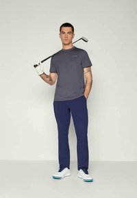 Šedé krátké sportovní tričko s rukávy, modré golfové kalhoty, bílé golfové boty, držící golfovou hůl s bílou rukavicí, stojící na jednobarevném pozadí.