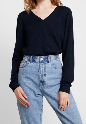 Marinblå långärmad V-ringad topp i mjukt material, i kombination med högmidjade ljusblå denimjeans med knäppning och fickor.