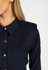 Vrouw met een marineblauwe ribbelige button-up shirt met een puntige kraag en kleine stud oorbel op een effen achtergrond.