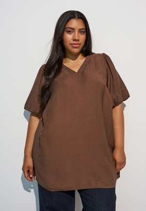 Donna con lunghi capelli scuri che indossa una maglia marrone larga con scollo a V e maniche corte a sbuffo e jeans blu scuro, in piedi contro uno sfondo semplice.