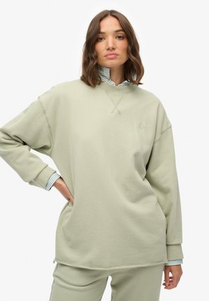 Sudadera oversize de color verde claro con un cuello acanalado, que presenta un logo sutil y un acabado texturizado. Combinada con una camisa a rayas debajo.