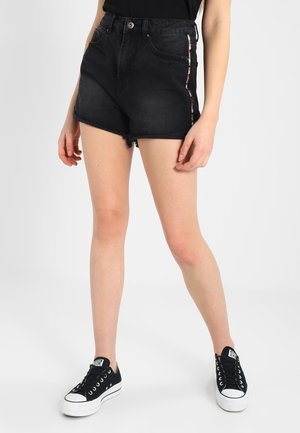 Bermudas de mezclilla negras con cintura alta, dobladillo deshilachado y rayas multicolores a lo largo del costado. Combinadas con zapatillas negras para crear contraste.
