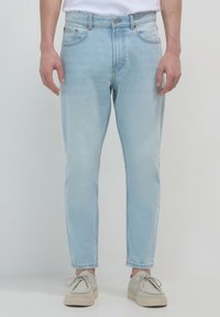 Uomo che indossa jeans slim-fit blu chiaro e scarpe stringate di colore bianco sporco, in piedi contro uno sfondo luminoso e semplice.