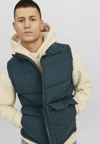 Dark green puffer vest with a zippered front, standing collar, and two side pockets, layered over a light beige hoodie.