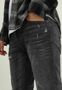 Los jeans negros desgastados presentan un desvanecimiento sutil y desgarros, detalles de costura y un ajuste slim. El modelo lleva una camisa de cuadros sobre una camiseta blanca.