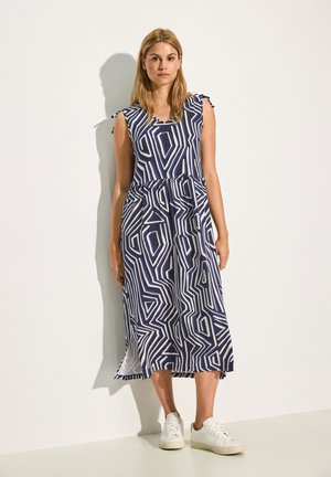 Navy- und weiße geometrisch gemusterte Kleid mit V-Ausschnitt, kurzen Ärmeln und seitlichen Schlitzern, kombiniert mit weißen Sneakers.