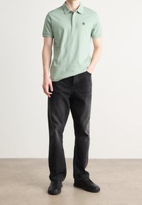 Camisa polo verde claro con cuello, mangas cortas y un parche de logo. Combinada con jeans negros de pierna recta y zapatillas negras.