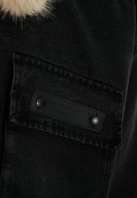 Chaqueta de mezclilla negra con un cuello de piel, con un tejido texturizado, dos botones de presión negros y una etiqueta en el bolsillo frontal.