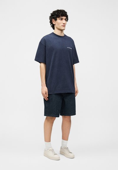 Un jeune homme se tient debout portant un T-shirt oversize bleu marine foncé, un short noir à fines rayures, des chaussettes blanches et des baskets beiges sur un fond clair uni.
