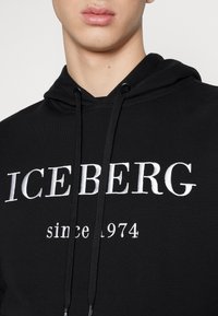 Schwarzer Hoodie mit weißem, gesticktem "ICEBERG"-Logo und dem Text "seit 1974". Verstellbare Zugbänder am Kragen. Glatte Stoffoberfläche.