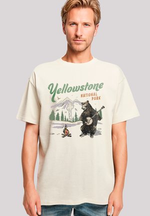 Beige cotton t-shirt featuring a graphic of a bear playing a banjo, with "Yellowstone National Park" text, surrounded by mountains and trees.