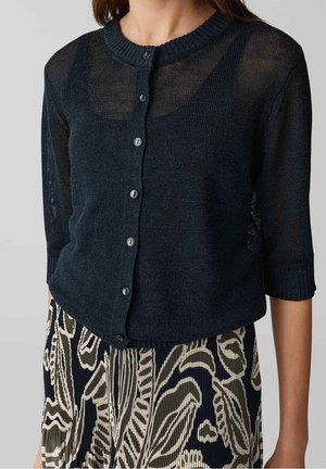 Mujer que lleva un cárdigan oscuro azul marino transparente abotonado sobre una camiseta de tirantes negra y una falda plisada con un patrón abstracto en negro, blanco y oliva.