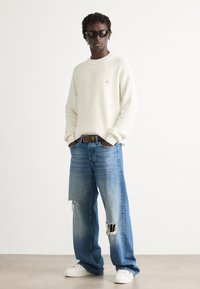 Man in een oversized witte trui, gescheurde wijde blauwe jeans, witte sneakers en een zonnebril, staand met zijn handen in zijn zakken op een effen achtergrond.
