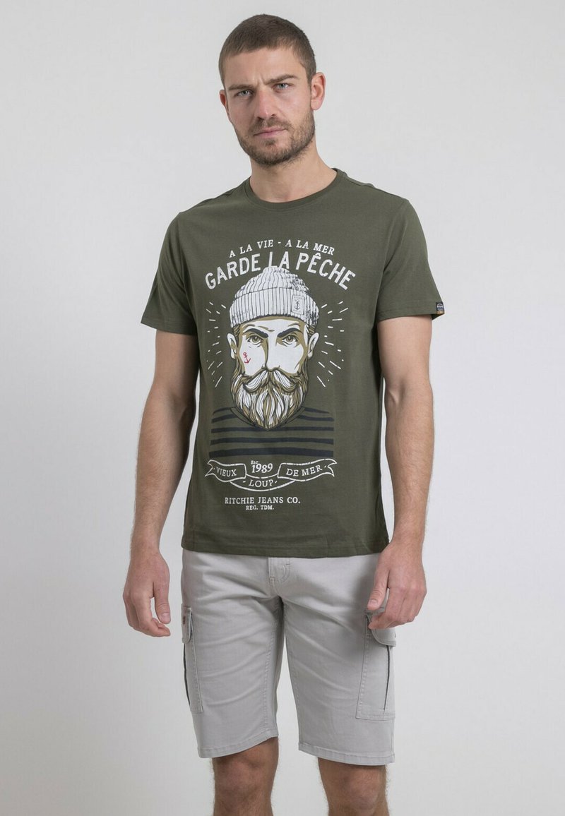 T-shirt en coton vert olive avec un design graphique d'un pêcheur barbu, avec du texte imprimé et des rayures horizontales en bas.