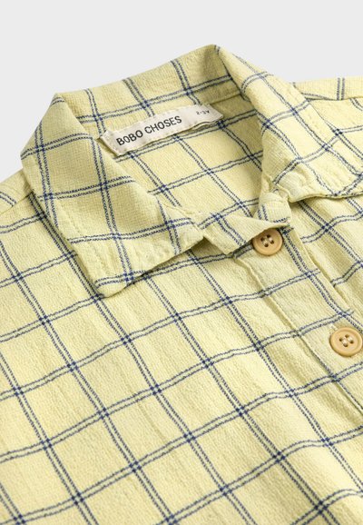 Chemise en tissu jaune texturé avec motif à carreaux bleus, boutons marron, et étiquette "BOBO CHOSES" à l'intérieur du col.