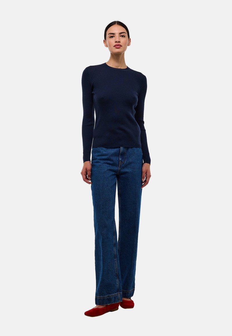 Haut à manches longues côtelé marine associé à un jean large à taille haute en denim bleu et des chaussures rouges. Design simple, texture lisse.