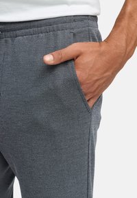 Grau strukturierter Jogginghose mit elastischem Bund und Seitentaschen. Eine Hand ist in die linke Tasche gesteckt, was den weichen Stoff und das legere Design zeigt.