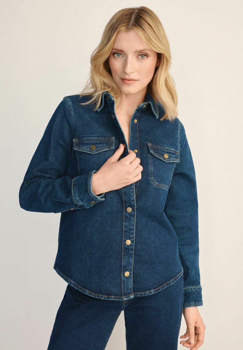 Niebieska koszula dżinsowa w ciemnym odcieniu, z zapięciem na guziki z przodu, dwoma kieszeniami na piersi i zaokrąglonym dołem. Tekstura materiału jest gładka z widocznymi szwami.