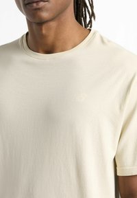 Cream short-sleeve t-shirt made of soft fabric, featuring a crew neckline, subtle branding on the chest, and textured detailing on the sleeves.