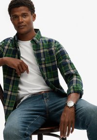 Grün-karierte und marineblaue Flanellhemd über einem weißen T-Shirt, kombiniert mit blauen Jeans. Mit einer Uhr und einem braunen Gürtel. Stuhl sichtbar.