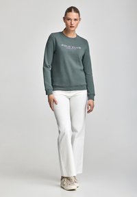 Sudadera verde con cuello redondo y gráfico de "POLO CLUB". Combinada con pantalones de campana blancos y zapatillas beige. Textura de tela suave.