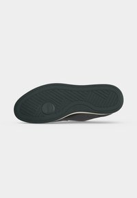 Semelle de chaussure en caoutchouc noir avec motif texturé antidérapant et détail de marque circulaire, vue par le dessous sur fond blanc.