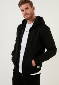 Crna zip-up dukserica s podstavljenim kapuljačom, dva prednja džepa i rebrastim manžetama. Nosila se preko bijelog T-shirt-a s otisnutom porukom.