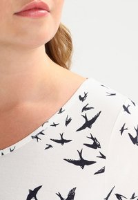 Top bianco con motivo di uccelli neri, con scollatura ampia e tessuto liscio, evidenziando un primo piano della zona della spalla.