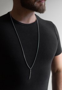 Silberne Kettenhalskette mit einem geschwungenen Anhänger, der an einer langen, dünnen Kette hängt. Das Modell trägt ein eng anliegendes schwarzes T-Shirt, was den Schmuck betont.