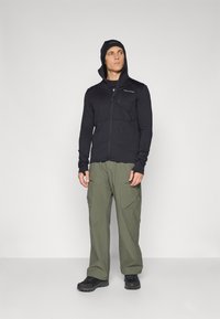 Giacca nera zip-up in tessuto testurizzato con cappuccio, abbinata a pantaloni cargo verde oliva con tasche laterali e una vestibilità rilassata. Scarpe nere.
