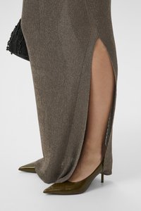 Detailní záběr nohy, která má na sobě hnědou pletenou sukni s vysokým rozparkem, v kombinaci s olivově zelenými lodičkami s špičatou špičkou a drží černou korálkovou kabelku.