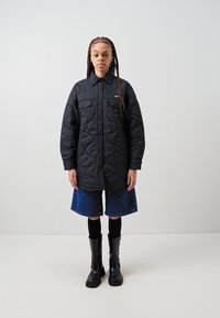 Black quilted jacket with a button-up front, two chest pockets, and a small logo. Worn with denim shorts and black knee-high boots.