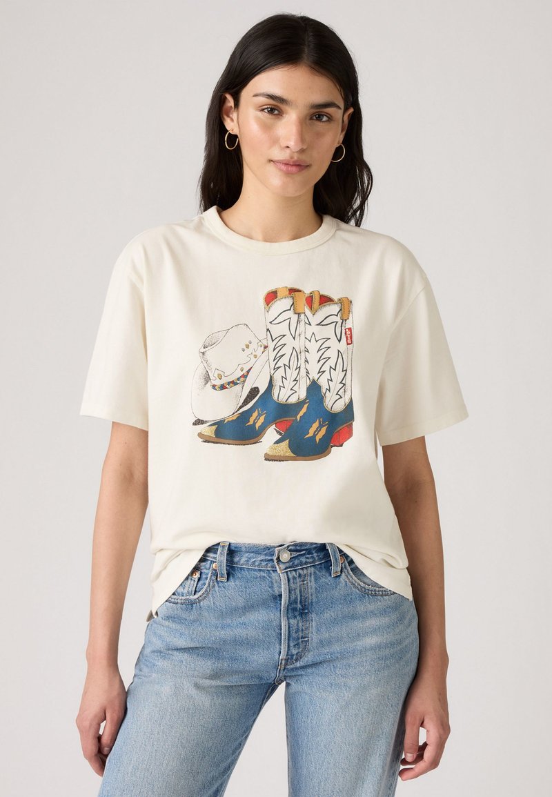 T-shirt crème arborant un graphique de bottes de cowboy et d'un chapeau. Les bottes sont bleues avec des accents jaunes ; le chapeau est blanc avec des détails colorés.