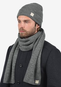 Pilka megzta kepurė ir šaliko rinkinys, pagaminti iš minkštos, tekstūruotos medžiagos. Šalikėlyje yra etiketė, derinama su tamsia striuke.