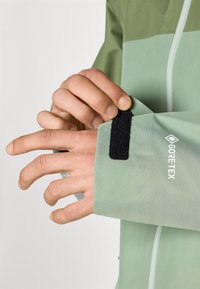 Veste GORE-TEX avec un schéma de couleurs vert clair et vert foncé ; comprend une fermeture à patte auto-agrippante au niveau des poignets et un tissu texturé.