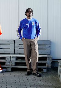 Blå Adidas-genser med italiensk flaggemblem, beige bukser og svarte sko. Står på en asfaltlagt overflate med trekasser i bakgrunnen.