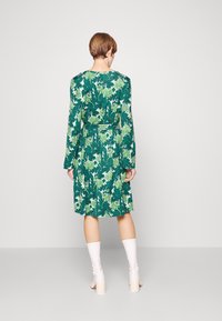 Abito floreale verde con vita cintrata, maniche lunghe e orlo a ginocchio. Presenta una combinazione di motivi verdi e bianchi. Indossato con stivali bianchi fino al ginocchio.