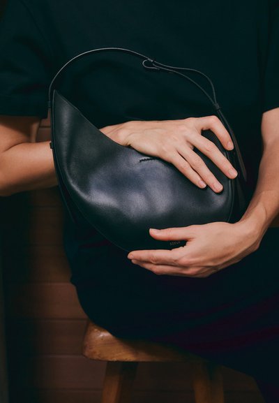 Sac à main en cuir noir avec une forme en croissant unique. Il présente un design minimaliste, une texture lisse et un logo discret. Une bandoulière réglable.