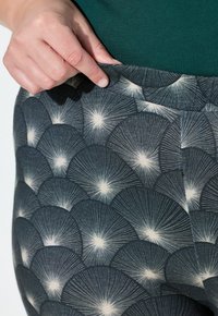 Patronen leggings in donkerblauw met crèmekleurige waaierontwerpen en stralende lijnen. Hoge tailleband wordt vastgehouden door een hand nabij de bovenrand.