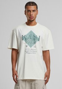 Kremfarget oversized T-skjorte med frontgrafikk i teal som viser stilisert tekst og en diamanform. Myk bomullsstoff, avslappet passform.